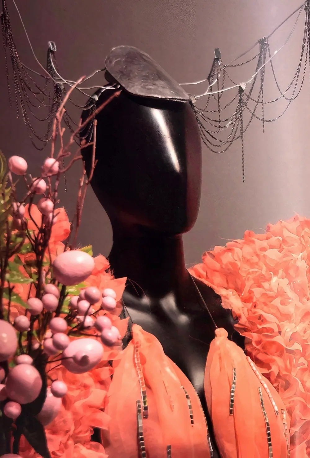 Buste de mannequin noir et brillant portant le costume rose à grands volants, le haut à paillettes et la coiffe de chaînes, à côté de branches et de baies roses.