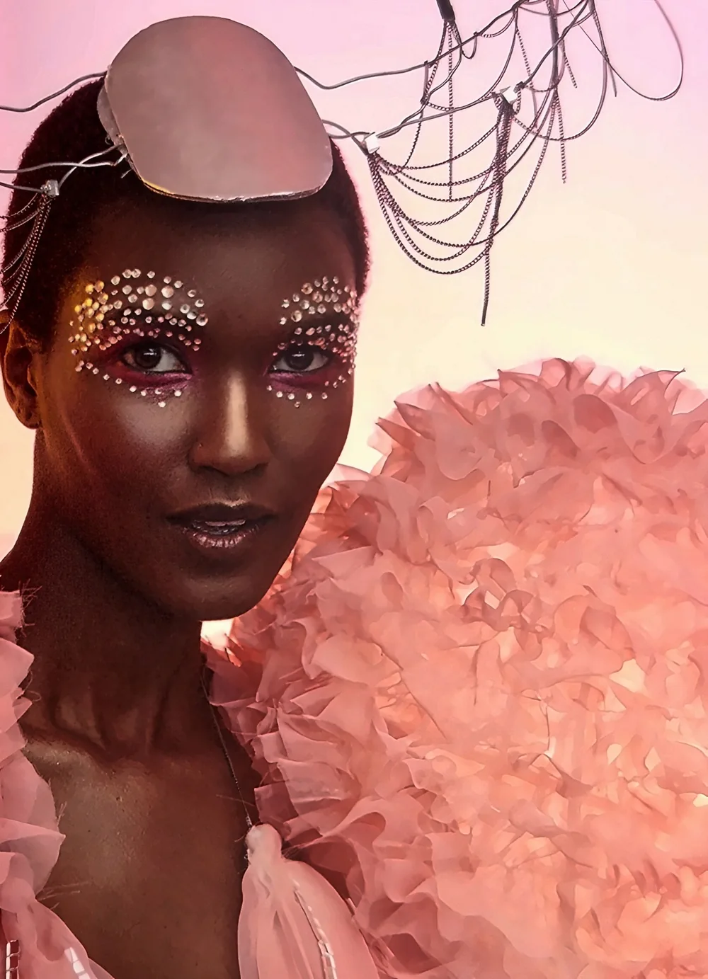 Portrait d'une mannequin noire au maquillage de strass, coiffe de chaînes avec plaque ovale rose et épaule en volants d'organza rose, sur fond rose.