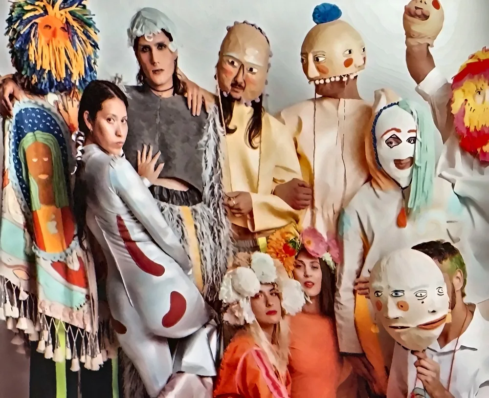 Groupe de performeurs en costumes colorés d'art portable, masques peints, coiffes en laine et en fleurs, sur fond clair.