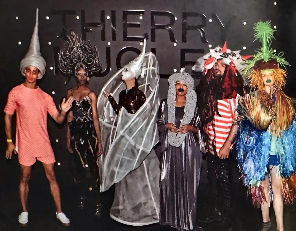Six mannequins en costumes d'art portable extravagants posant devant le lettrage THIERRY MUGLER sur un mur sombre constellé de points lumineux.