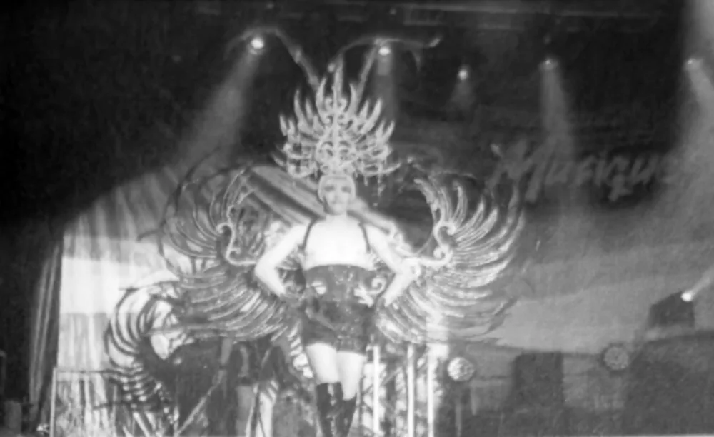 Noir et blanc : interprète de face en costume de plumes, haute coiffe rayonnante et grandes ailes déployées, sous les projecteurs.
