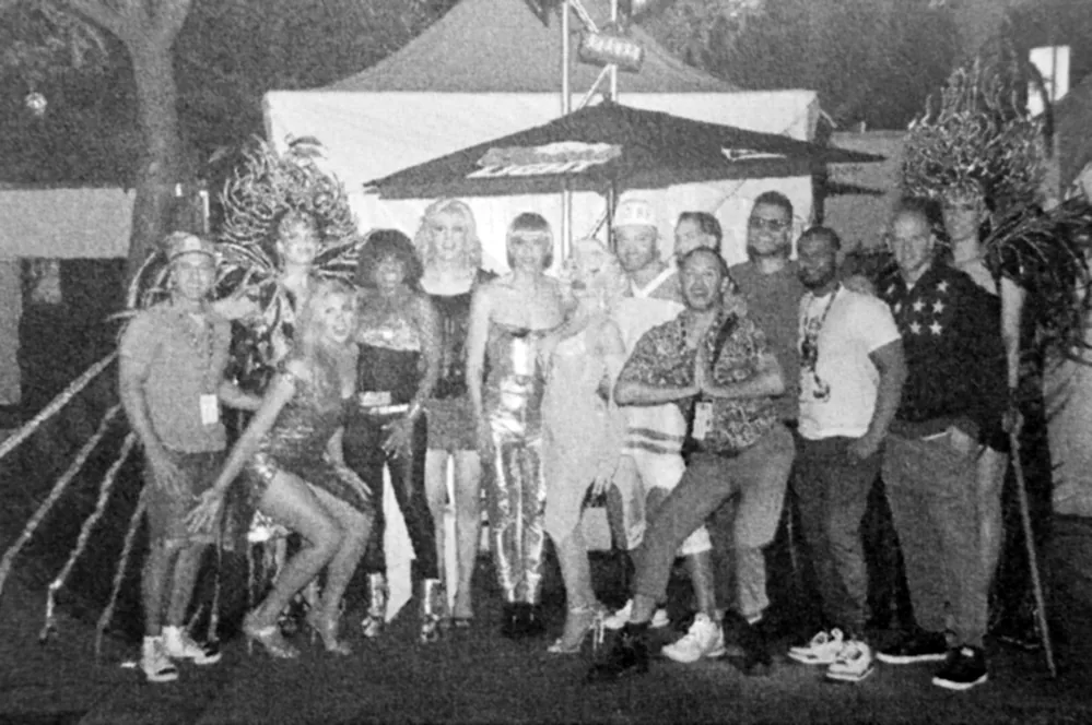 Noir et blanc : une quinzaine de personnes posent devant une tente et un parasol, coiffes de plumes aux deux extrémités du groupe.